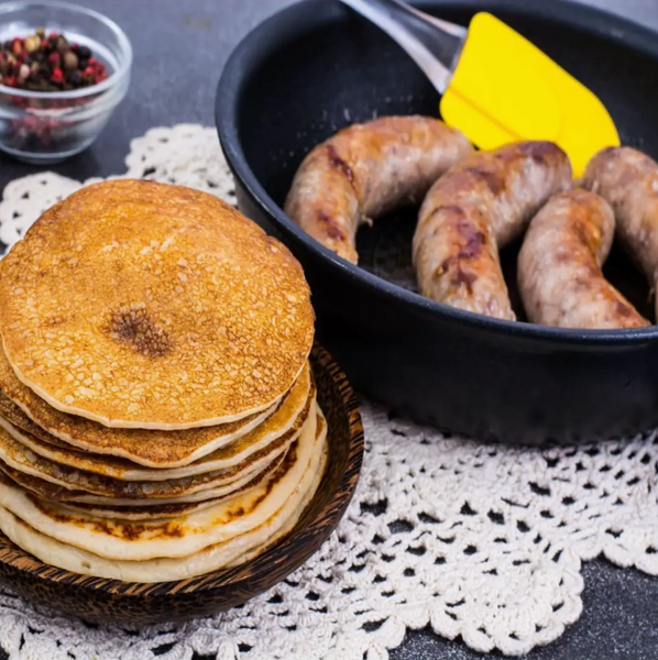 Stack of pancakes next to a pan with sausages on a lace tablecloth.