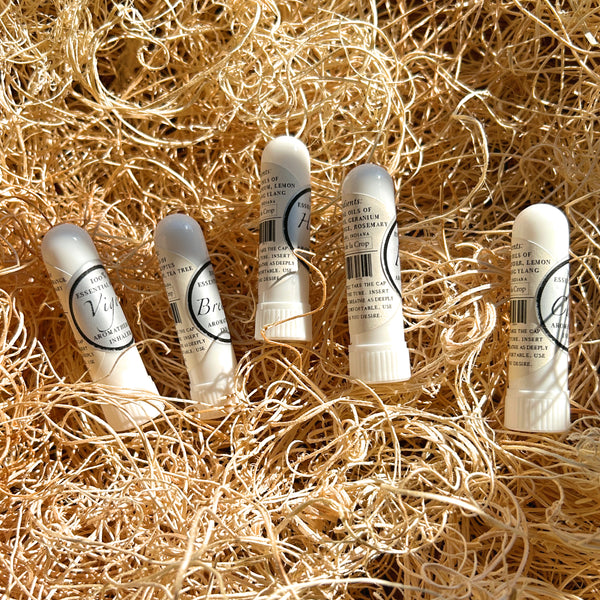 Five small white lip balm tubes with clear caps are arranged on a bed of shredded natural straw. The labels show different scents including Vanilla, Brown Sugar, Lemon, Mint, and Rosemary.
